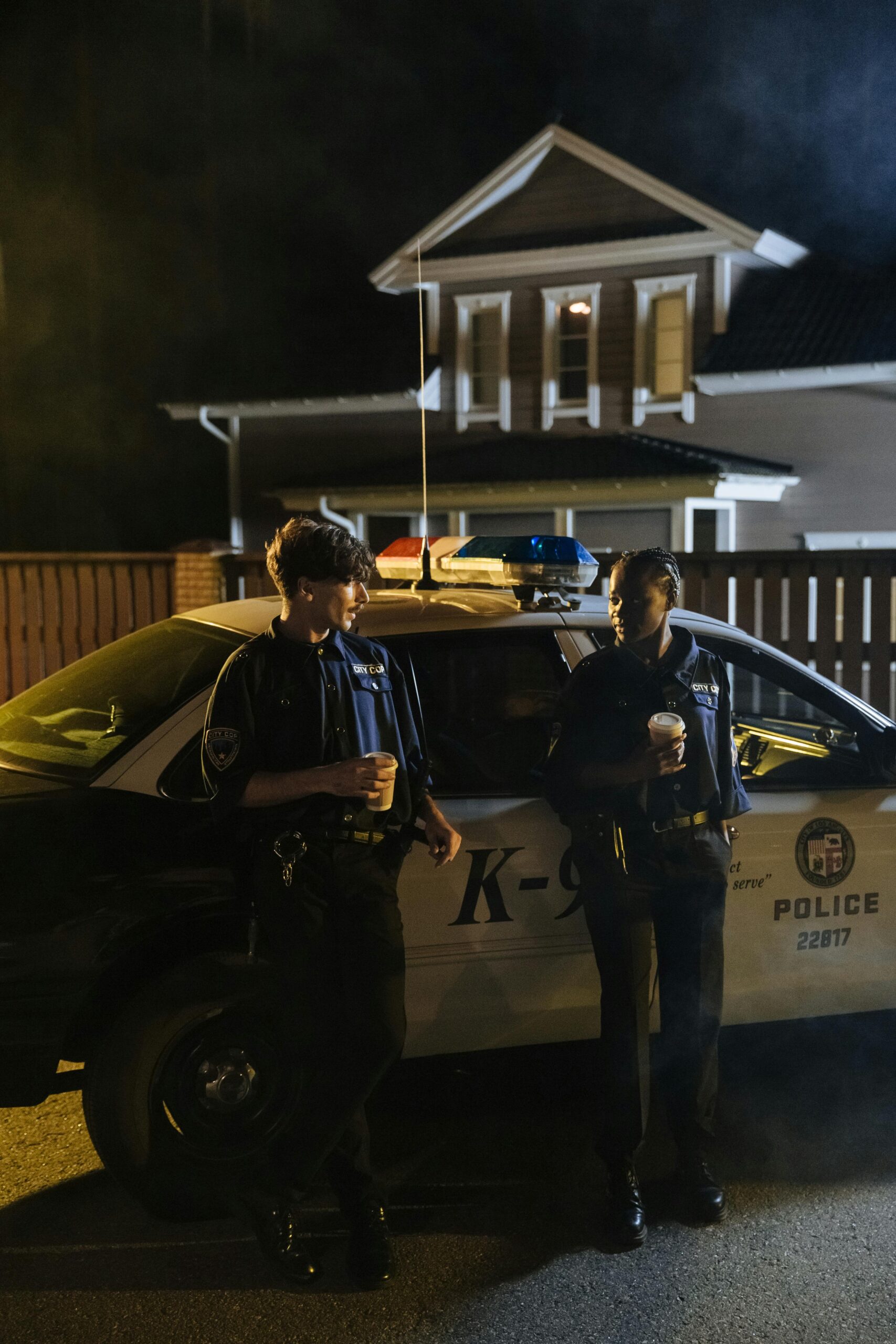 Two officers stand beside their police car during a night shift, holding coffee.